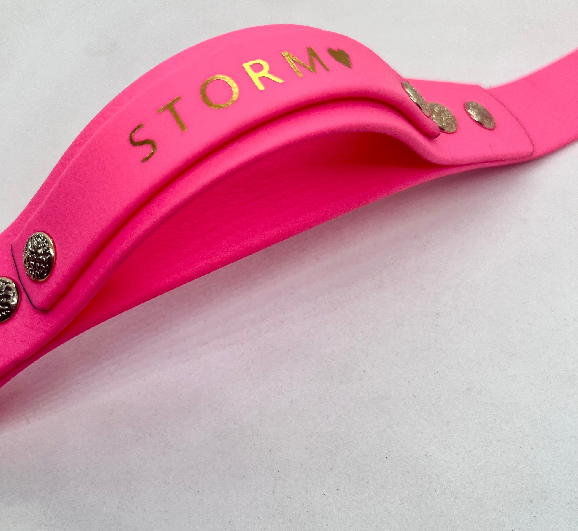 A close-up of a bright neon pink dog collar, featuring the name 'STORM' and a small heart foiled on the handle, with gold textured decorative rivets. JOOLS LONDON
