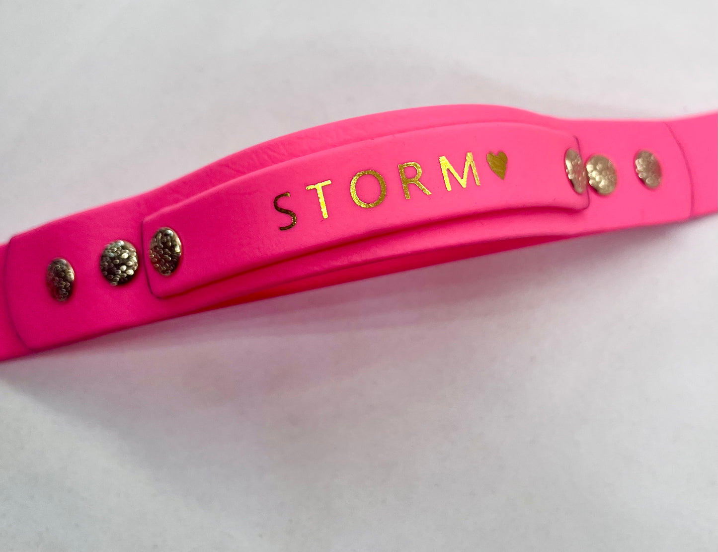 A bright pink beautifully finished collar with the name 'STORM' and a small heart in gold foil lettering, accented with gold textured studs on both ends, shown on a plain white background. JOOLS LONDON