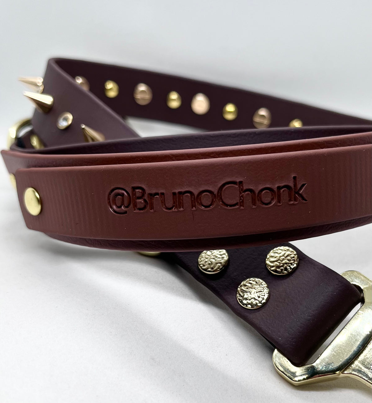 Close-up of the embossed @BrunoChonk handle on a brown BioThane® collar with gold-toned studs and buckle.