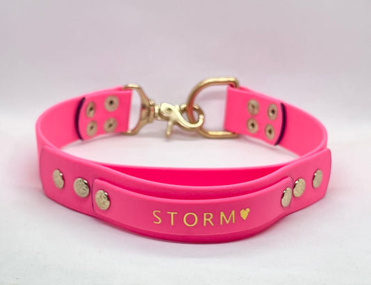 A bright pink pet collar with brass hardware, featuring the name 'STORM' and a small heart foil embossed on the handle, displayed on a light background.